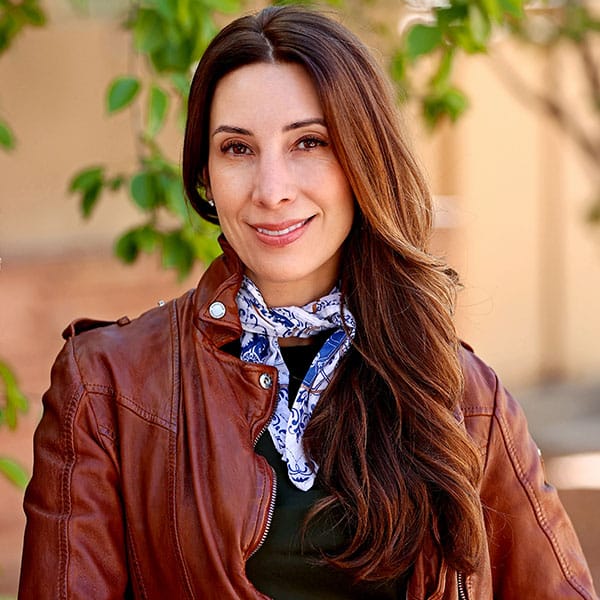 Marissa Ruyle with long brown hair wearing a brown leather jacket, a dark top, and a patterned scarf stands outdoors in front of green foliage and a beige wall, smiling at the camera.